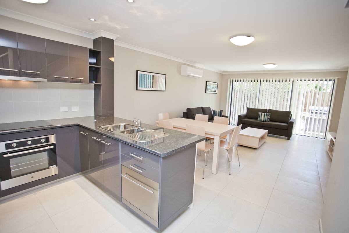 A modern kitchen with built-in appliances, a dining area with a table and chairs, and a living area with a sofa and a window.