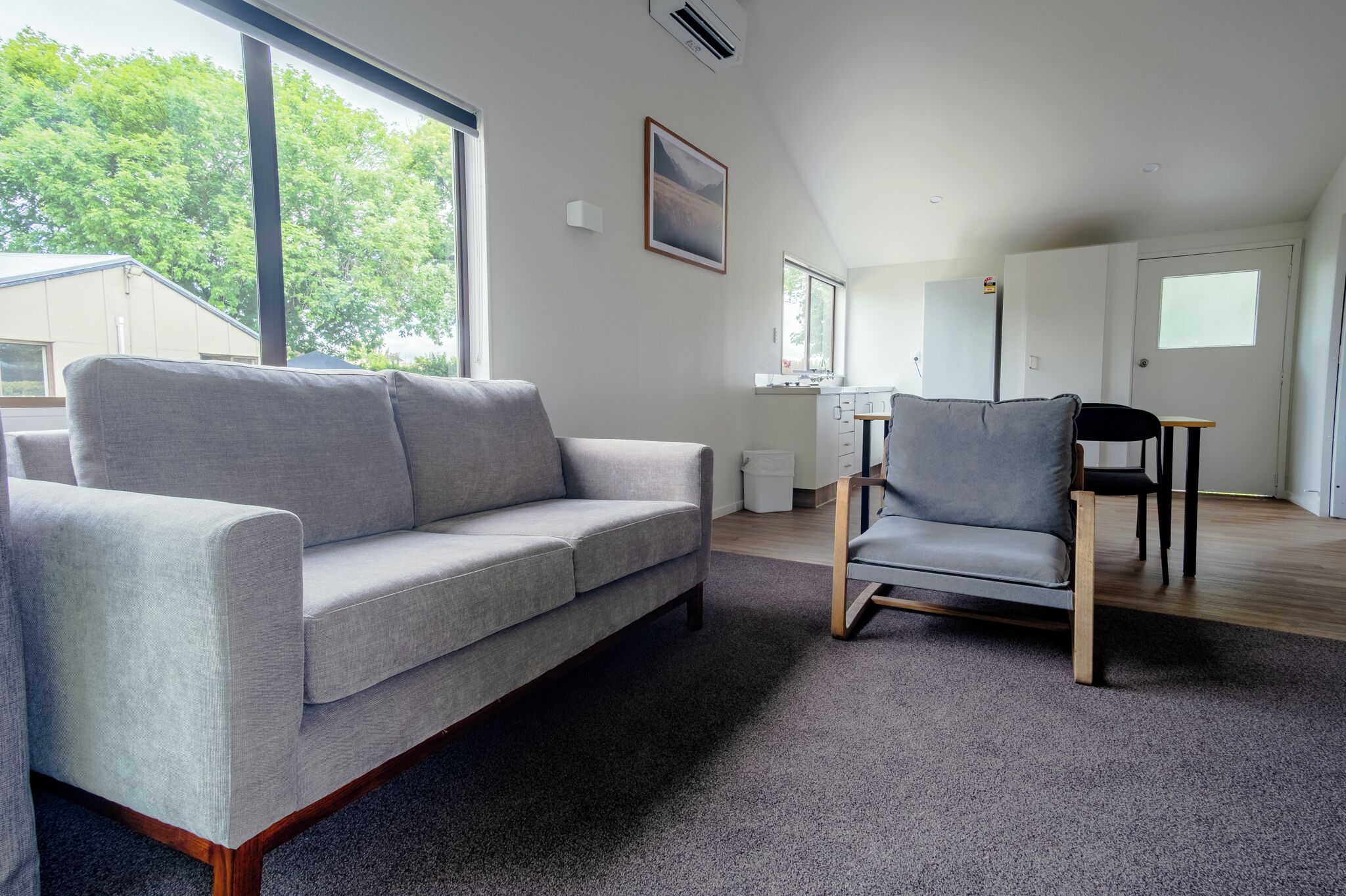 Un salon moderne avec un canapé gris, un fauteuil gris et une table à manger avec des chaises. Une grande fenêtre offre une vue sur les arbres à l’extérieur.