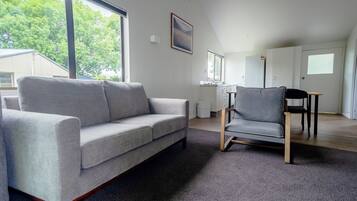 Un salon moderne avec un canapé gris, un fauteuil gris et une table à manger avec des chaises. Une grande fenêtre offre une vue sur les arbres à l’extérieur.