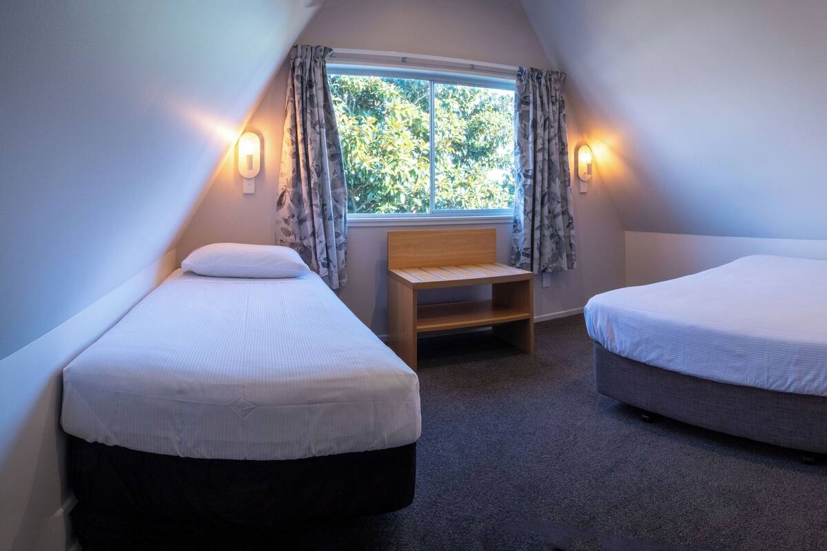 Een kleine, driehoekige kamer met twee eenpersoonsbedden, een houten bankje en een raam met gordijnen.
