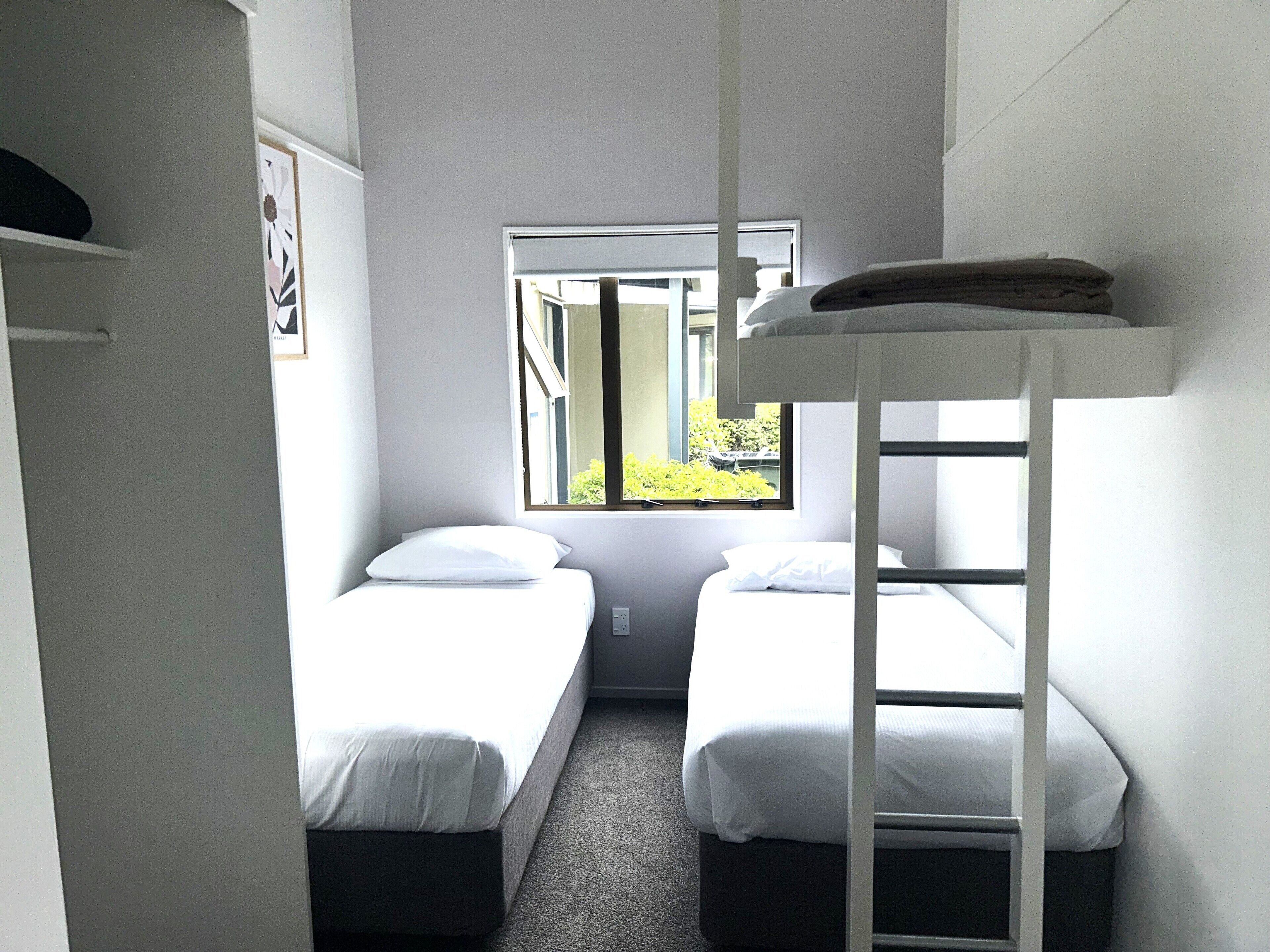Una habitación pequeña y limpia con dos camas individuales, una litera y una ventana con vistas a la vegetación, y una puerta a la izquierda.