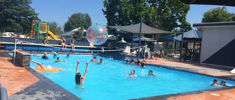 Una piscina con gente disfrutando del agua, una pelota inflable grande y un parque infantil al fondo.