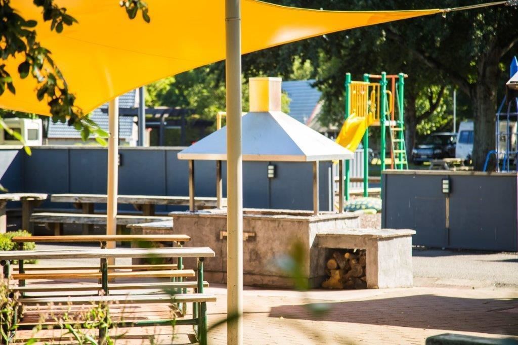 Un parque infantil con un tobogán amarillo y estructuras de escalada, bancos y una zona de estar cubierta.