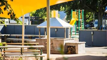 Un parque infantil con un tobogán amarillo y estructuras de escalada, bancos y una zona de estar cubierta.