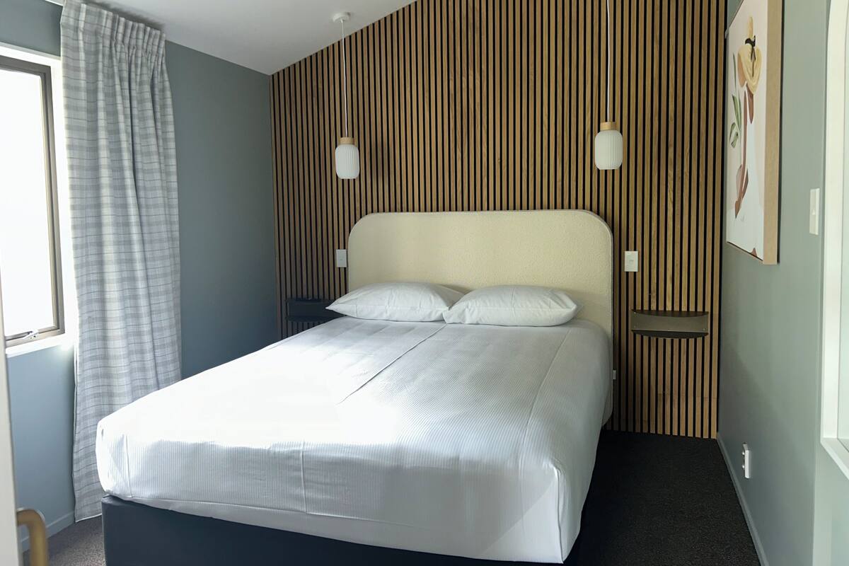 Een opgemaakt bed in een kamer met een gestreept behang, een raam met gordijnen en een wandlamp.