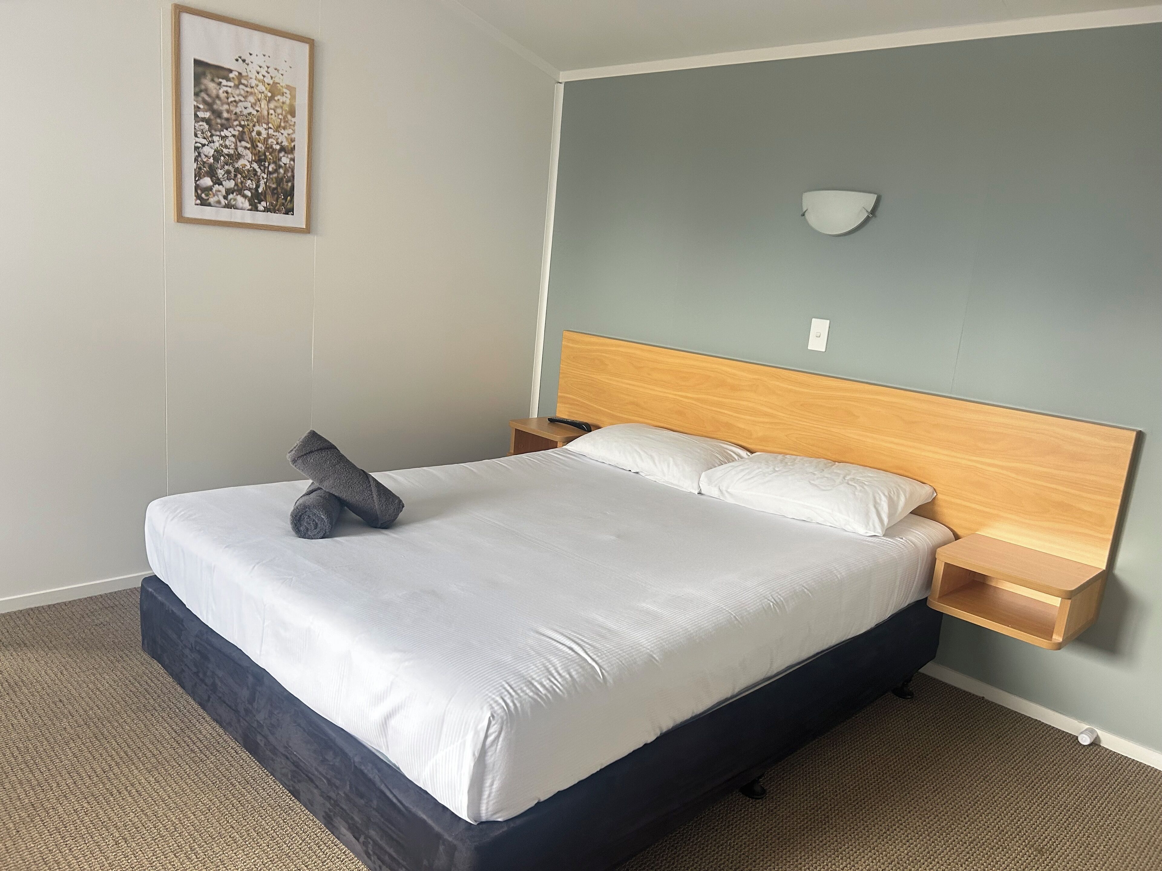 A neatly made bed with white linens and a dark headboard, a small bedside table with a lamp, and a framed picture on the wall.