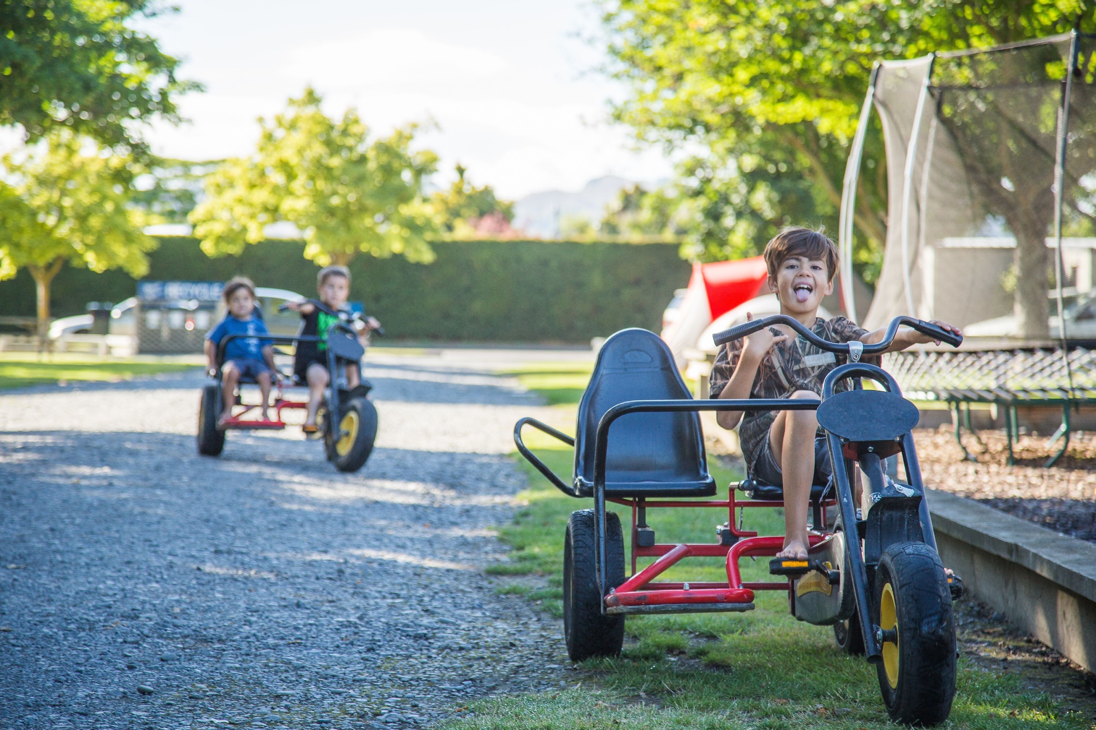 A gravel path with two children riding tricycles and a third child on a small cart.