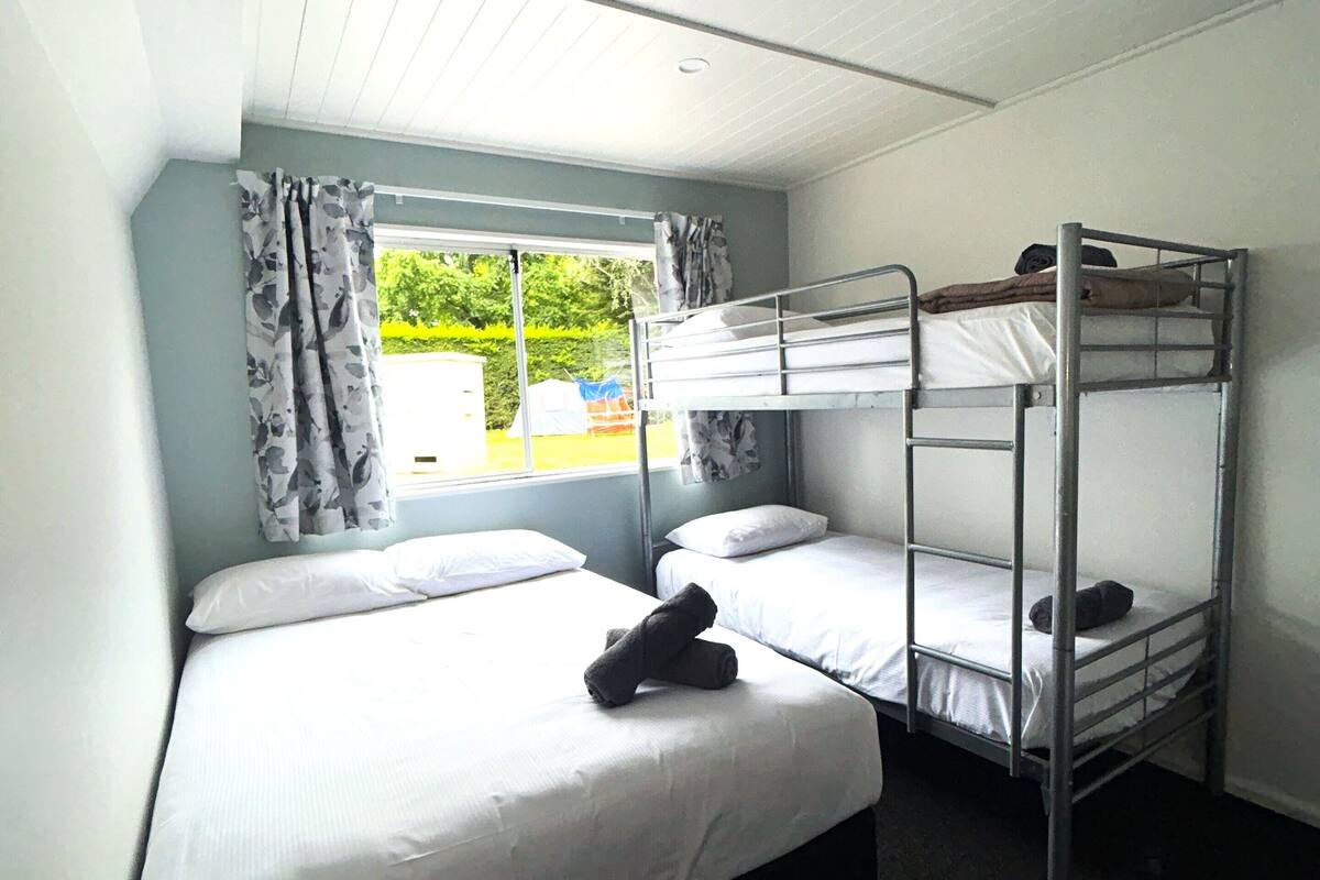 Een slaapkamer met stapelbed, raam, gordijnen en uitzicht op groen.