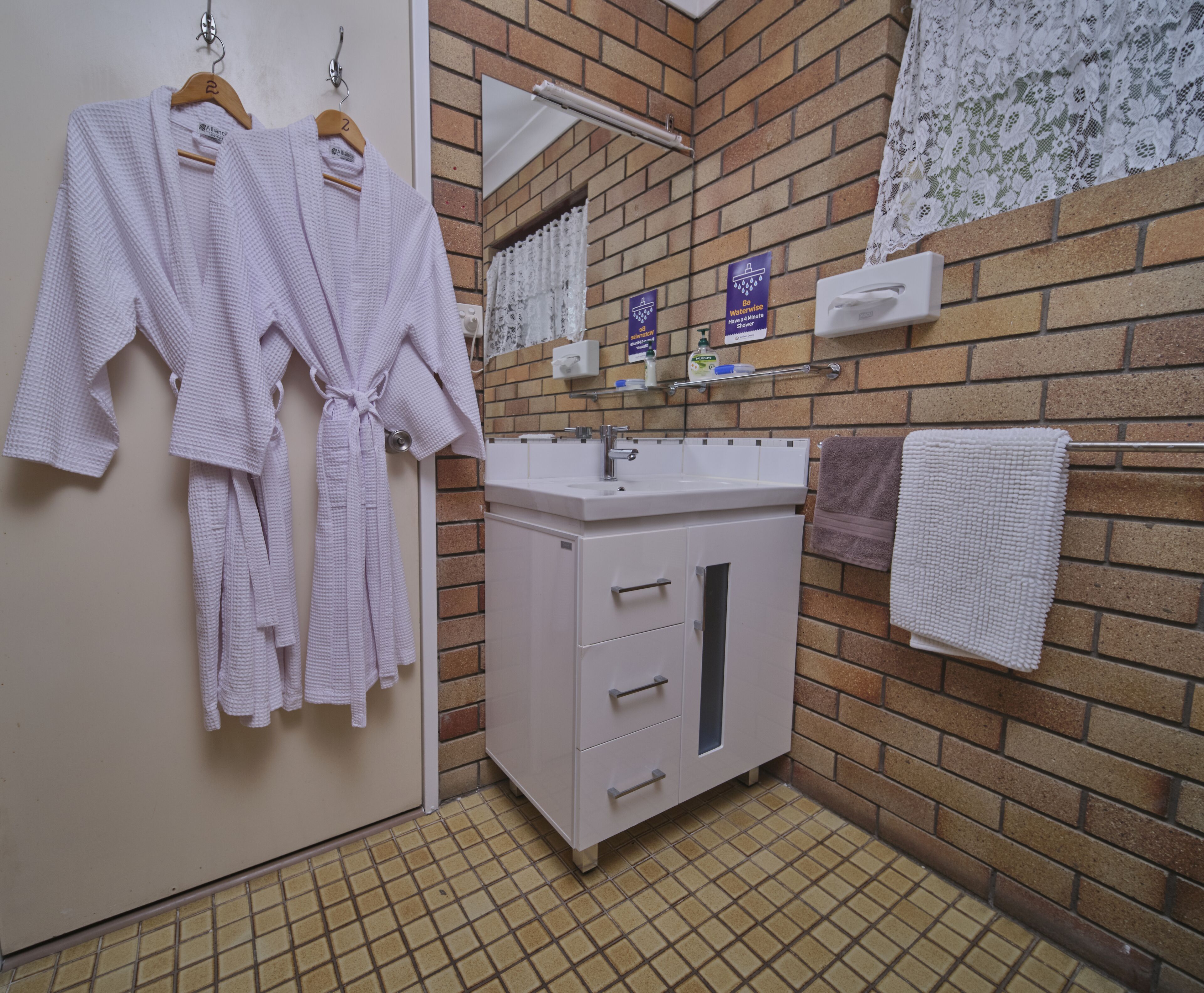 Un baño con lavamanos blanco, un espejo y un perchero. Hay dos batas blancas colgadas en un perchero de madera.