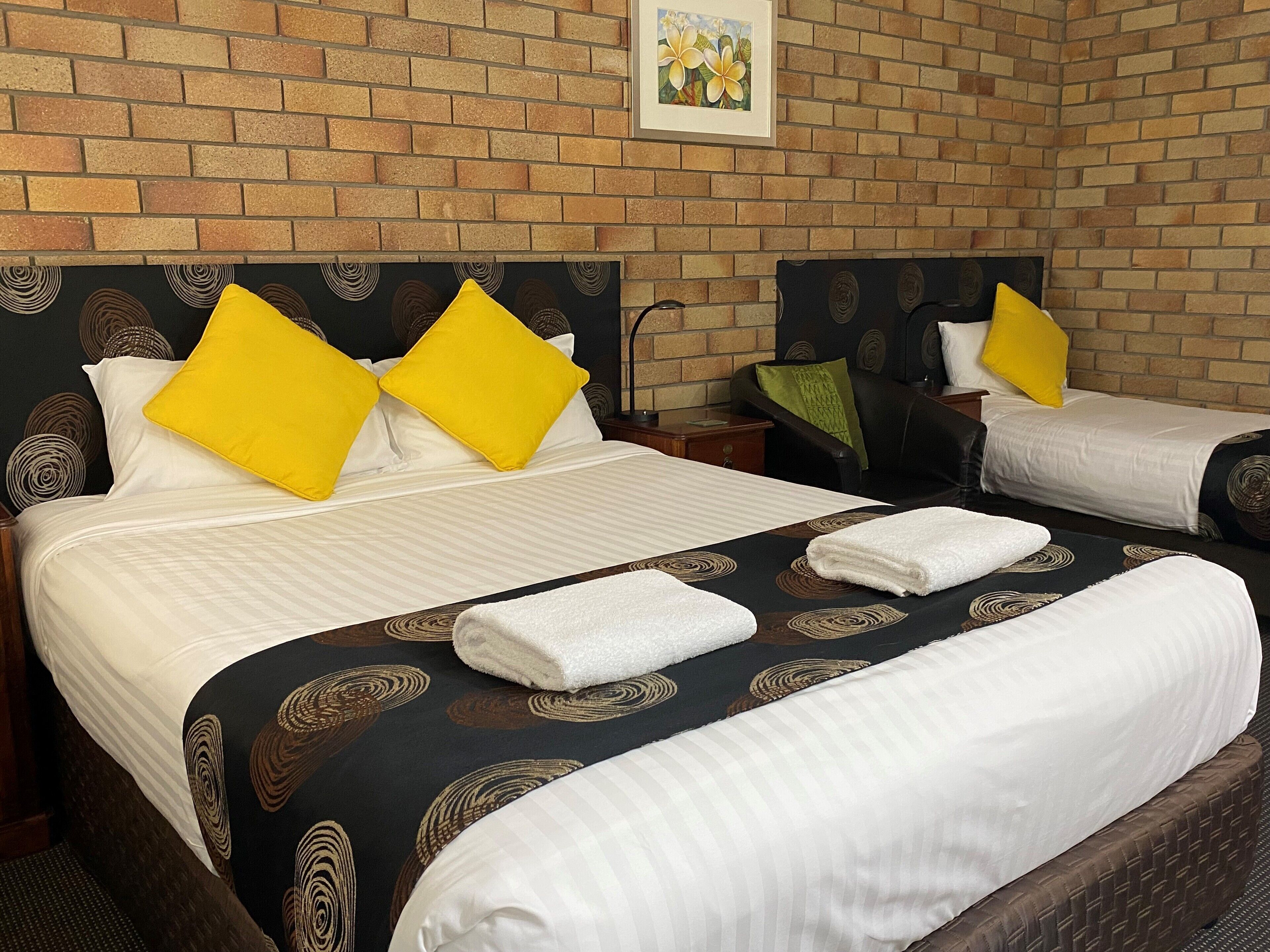 Ein Hotelzimmer mit zwei Betten, jeweils mit schwarzem Kopfteil und gelben Kissen. Das Zimmer verfügt über eine Ziegelwand und ein gerahmtes Bild über den Betten.