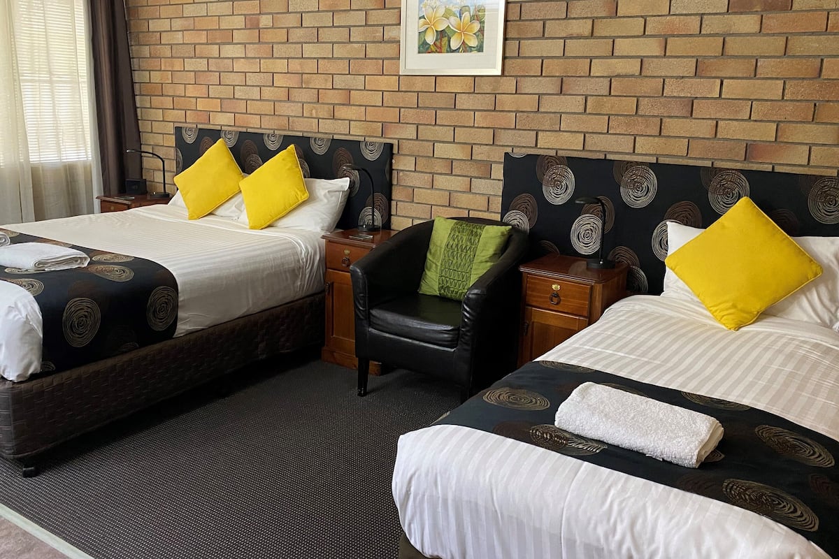 Habitación de hotel con dos camas, cada una con una colcha de diseño blanco y negro, un cojín verde y una almohada amarilla. Hay una butaca de cuero negro, una mesita de noche de madera con una lámpara y un cuadro enmarcado en la pared.