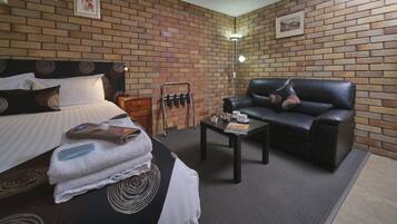 A hotel room with a brick wall, a bed with a headboard, a black leather sofa, a coffee table, and a small table with towels and a tea set.