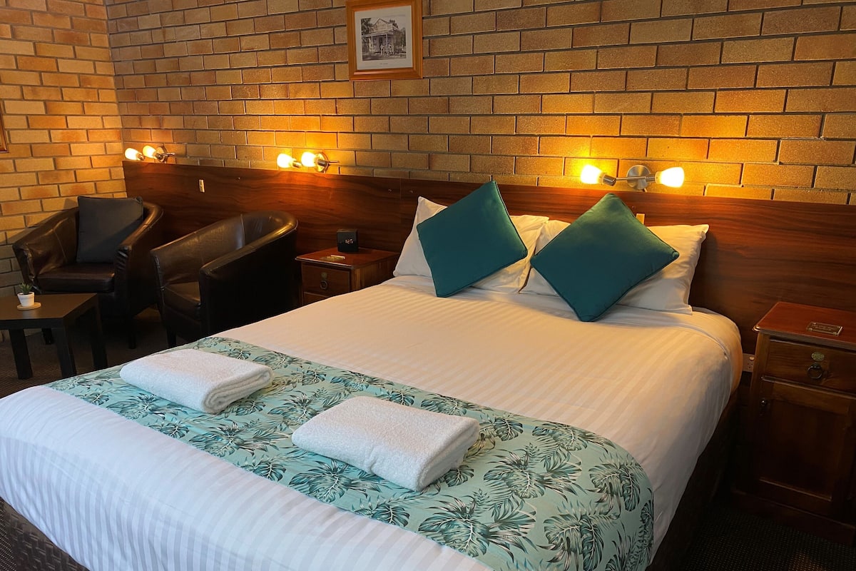Habitación de hotel con pared de ladrillo, cabecero de madera, cama con ropa blanca y almohadas con estampado de hojas verdes, mesita de noche de madera, sillón de cuero y una pequeña mesa con una planta.