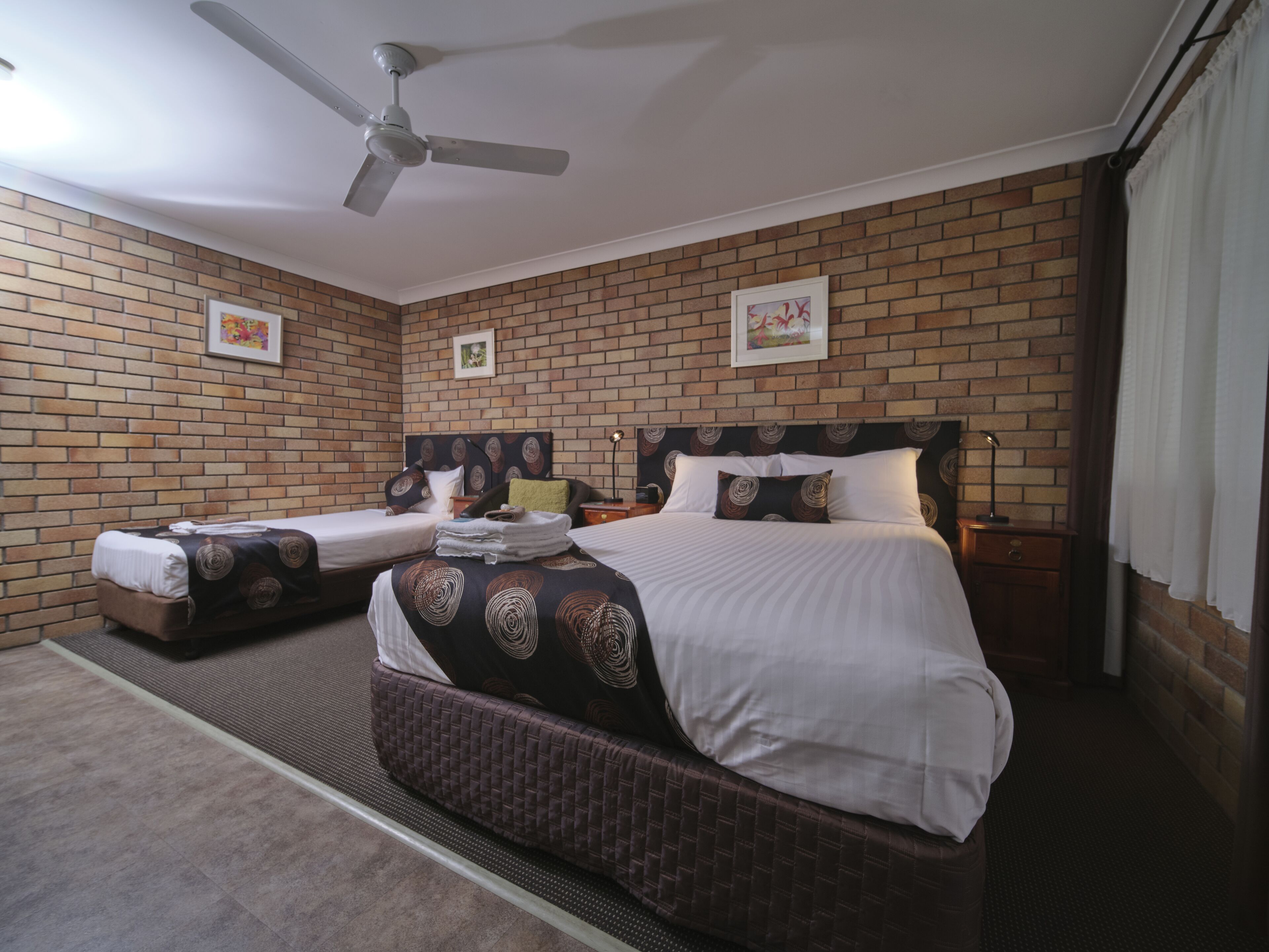 A hotel room with a brick wall, two beds, a ceiling fan, and artwork on the walls.