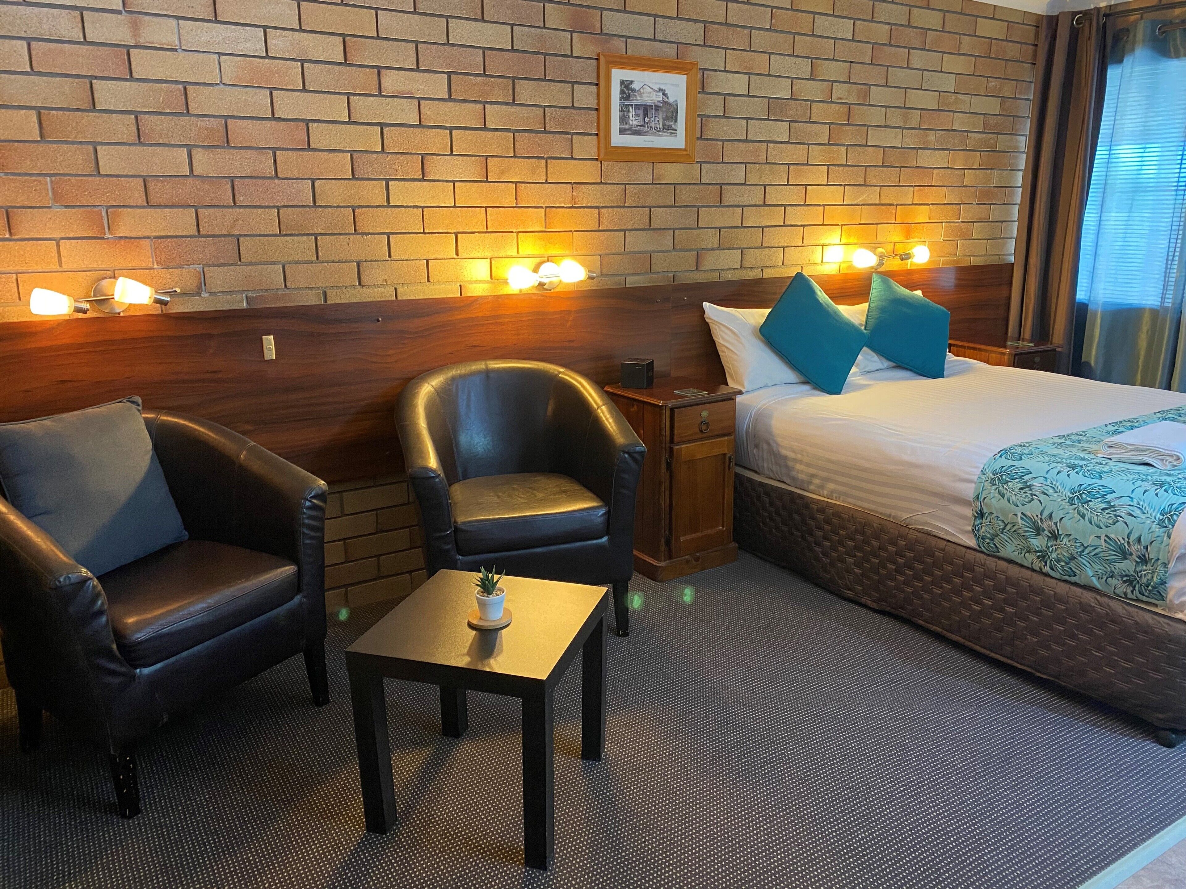 Habitación de hotel con pared de ladrillo, una cama con almohadas azules, una mesita con una planta y dos sillones de cuero.