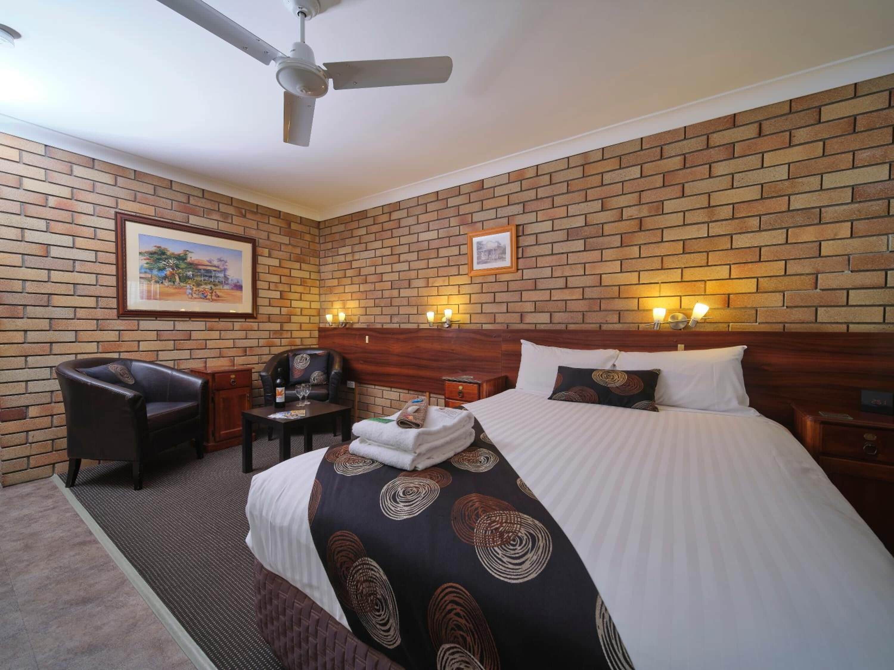A hotel room with a brick wall, a bed with a headboard, a desk with a chair, and a ceiling fan.