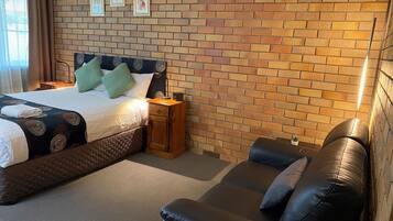 A hotel room with a brick-patterned wall, a bed with green and white bedding, a nightstand with a lamp, and a leather sofa.