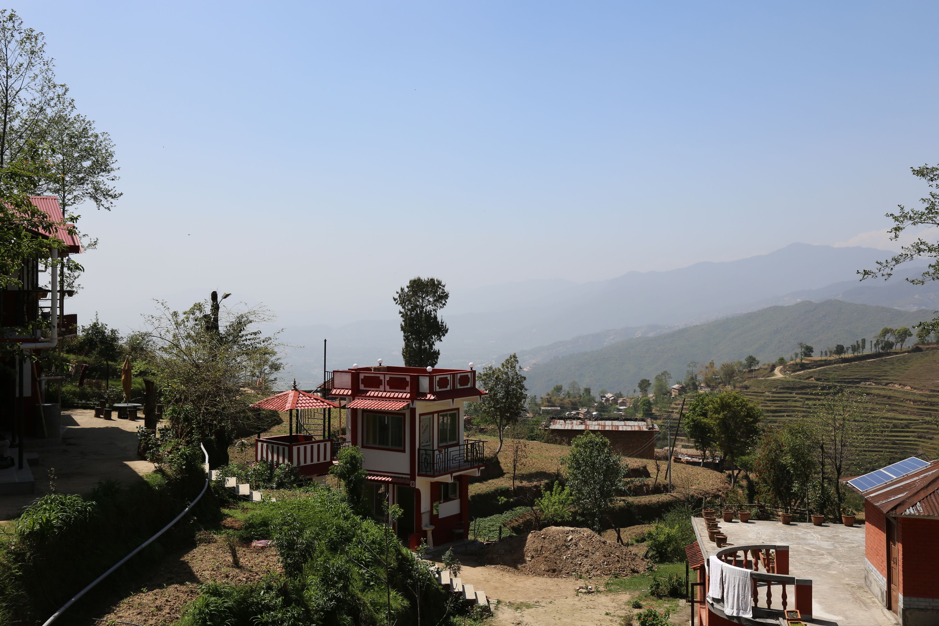 A two-story building with a red roof, surrounded by greenery and terraced fields, with mountains in the background.