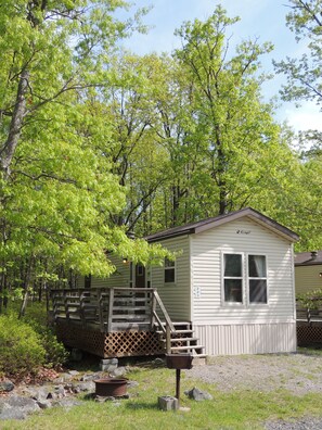 A small, single-story cabin with a wooden deck and a small porch, surrounded by lush green trees.