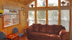 A cozy cabin interior with a wooden ceiling, large windows with blinds, a brown sofa, a small table, and two chairs.
