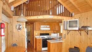 A wooden cabin interior with a kitchen, dining area, and a loft bedroom.