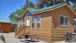 A wooden cabin with a green roof, a porch with a bench, and a trash can. - Oakzanita Springs RV Campground (Descanso)