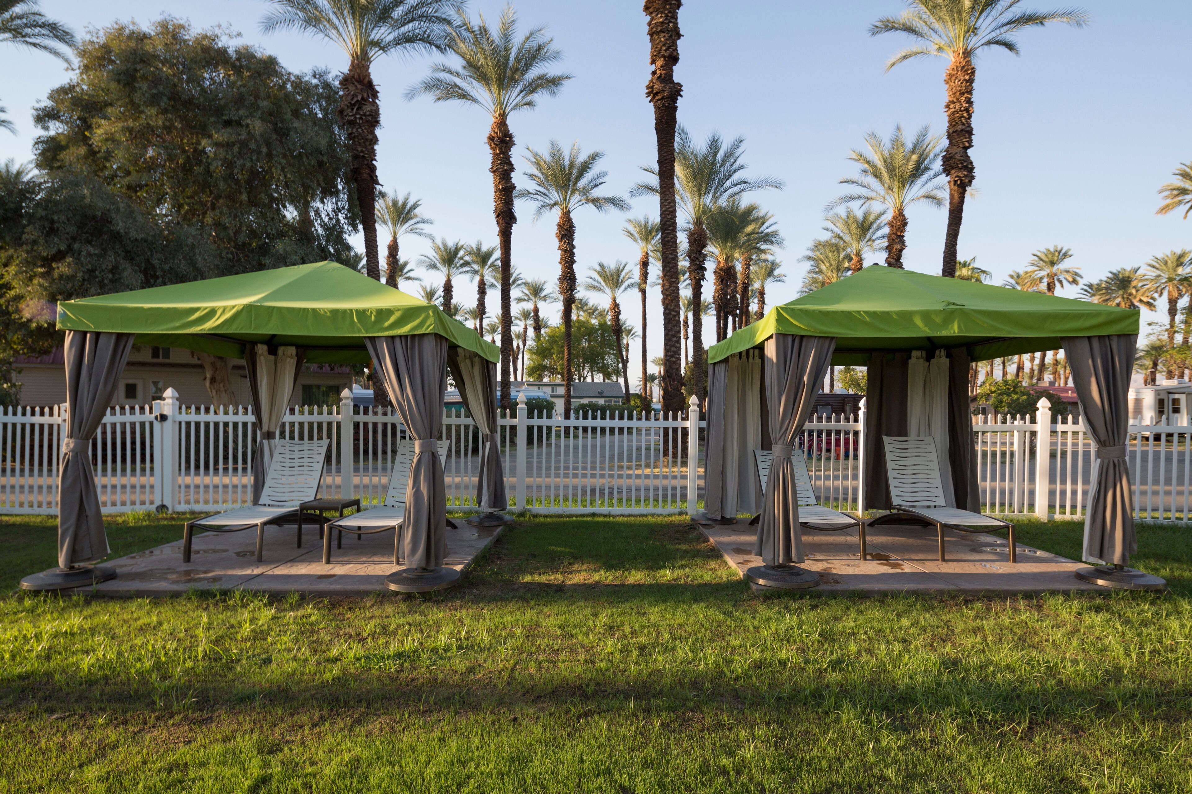 Two outdoor seating areas with green canopies, surrounded by palm trees and a white picket fence.