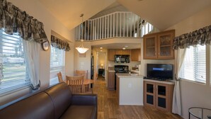 A spacious living area with a brown leather sofa, wooden dining table, and chairs. A kitchen with wooden cabinets and a built-in island is visible in the background. - Palm Springs RV Resort (Palm Desert)