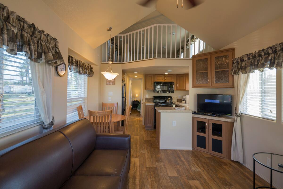 A spacious living area with a brown leather sofa, wooden dining table, and chairs. A kitchen with wooden cabinets and a built-in island is visible in the background.