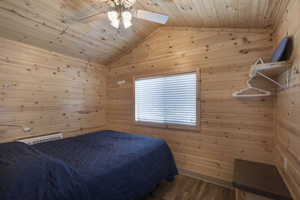 A small, cozy room with wooden walls, a single bed, a ceiling fan, a window with blinds, and a small shelf with a hanging item.