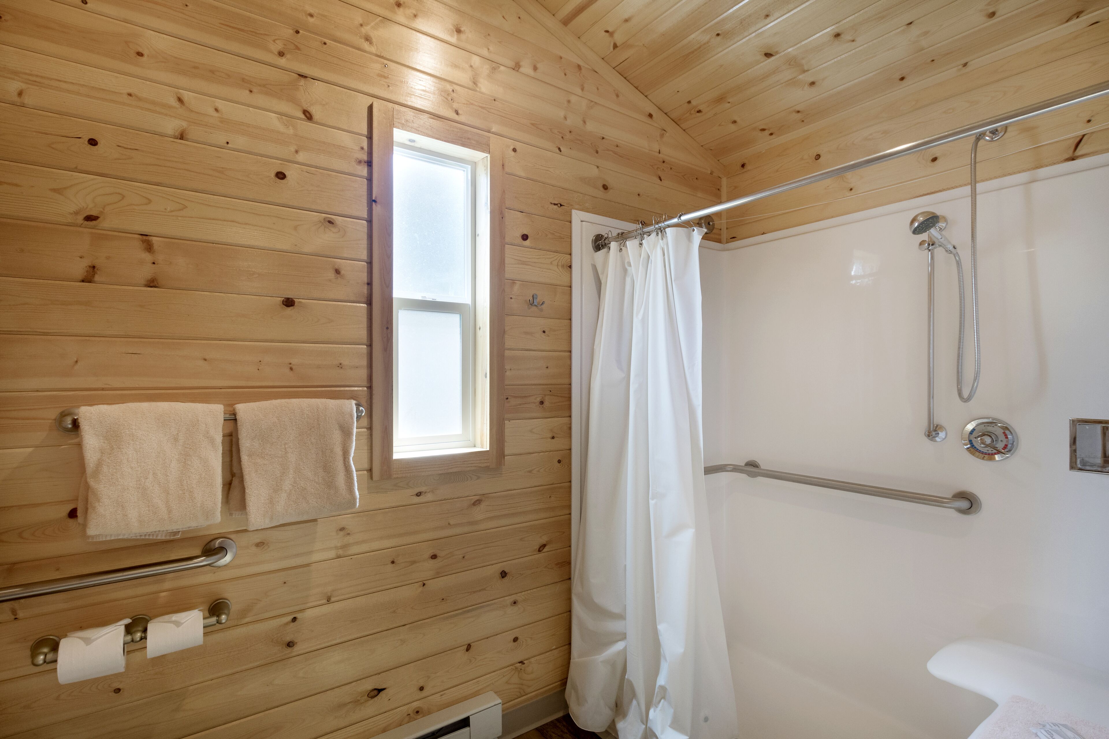 A wooden-paneled bathroom with a shower, grab bars, a window, and towels.
