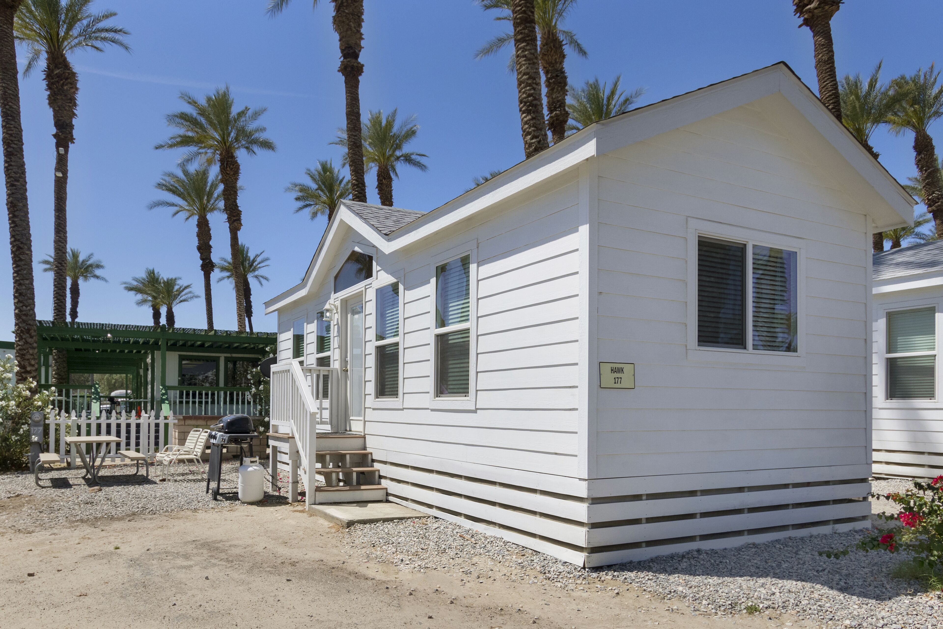 A small, single-story mobile home with a white exterior, numbered 177, surrounded by palm trees and a gravel driveway.