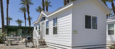 A small, single-story mobile home with a white exterior, numbered 177, surrounded by palm trees and a gravel driveway.