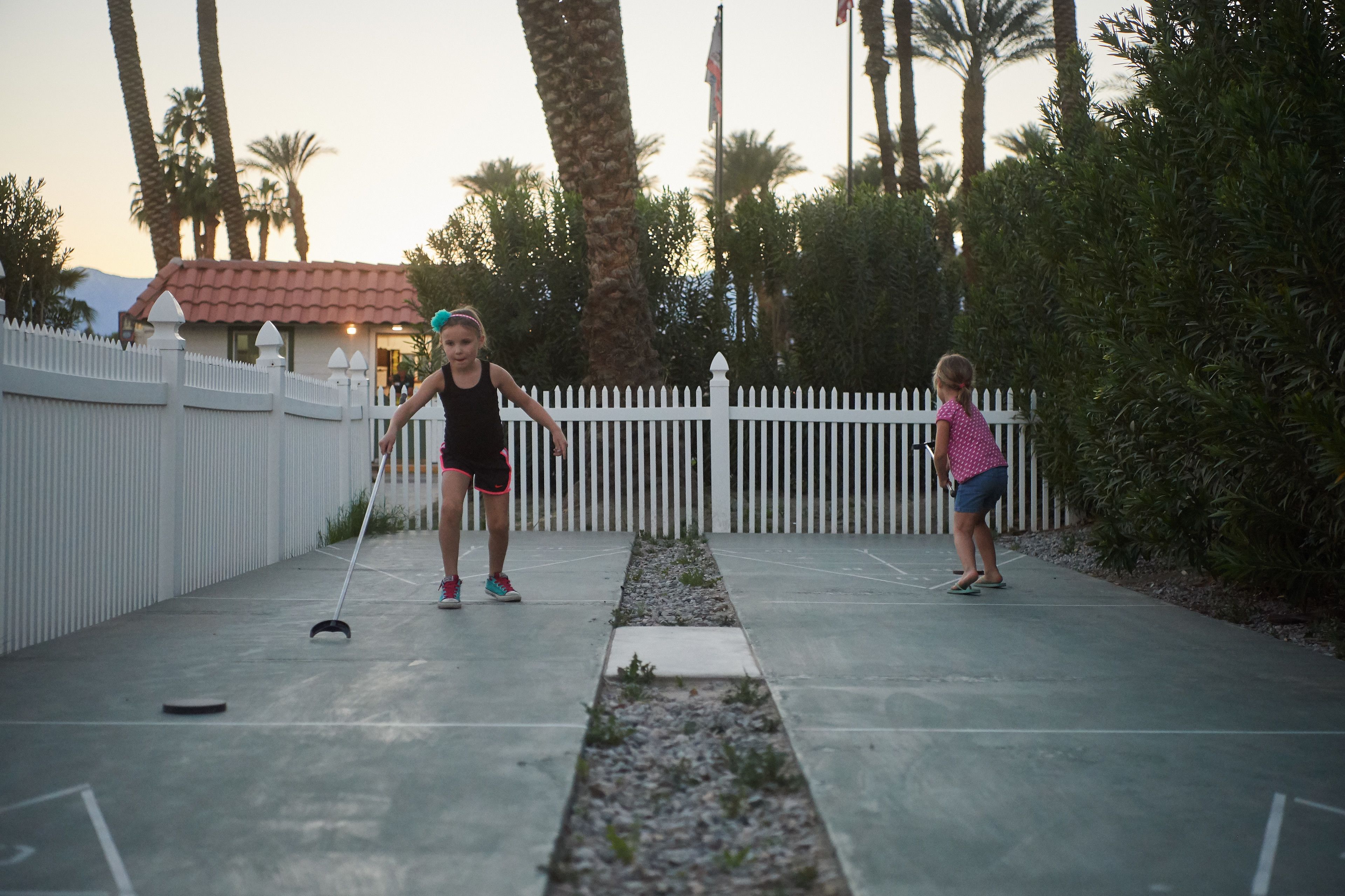 Two people playing shuffleboard on a concrete court with a white picket fence and palm trees in the background.