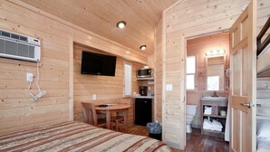 Quarto de cabana de madeira com cama, escrivaninha, televisão, micro-ondas e banheiro.