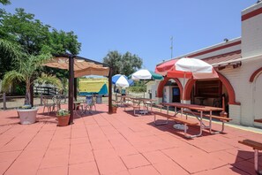 An outdoor patio area with picnic tables and umbrellas, surrounded by potted plants and a building with a red and white facade.