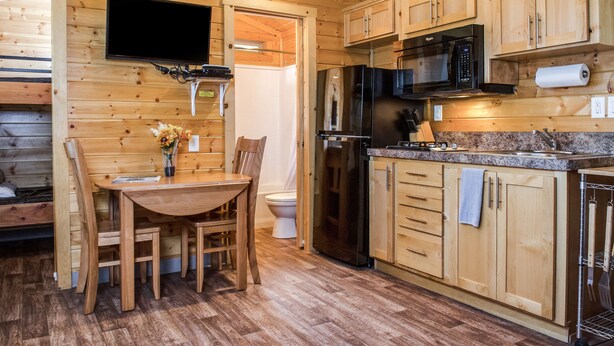 A wooden cabin interior with a dining table, chairs, a TV mounted on the wall, a refrigerator, a microwave, and wooden cabinets. - Russian River RV Campground (Cloverdale)
