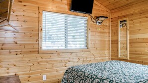 A wooden cabin room with a bed, a window with blinds, a mounted TV, and a wall-mounted light fixture. - Russian River RV Campground (Cloverdale)