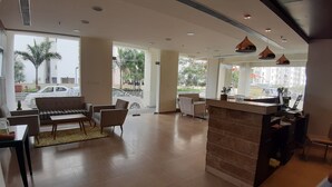 A spacious hotel lobby with a sofa, chairs, a reception desk, and large windows offering views of the street and palm trees. - Starlit Suites bangalore (Anekal)