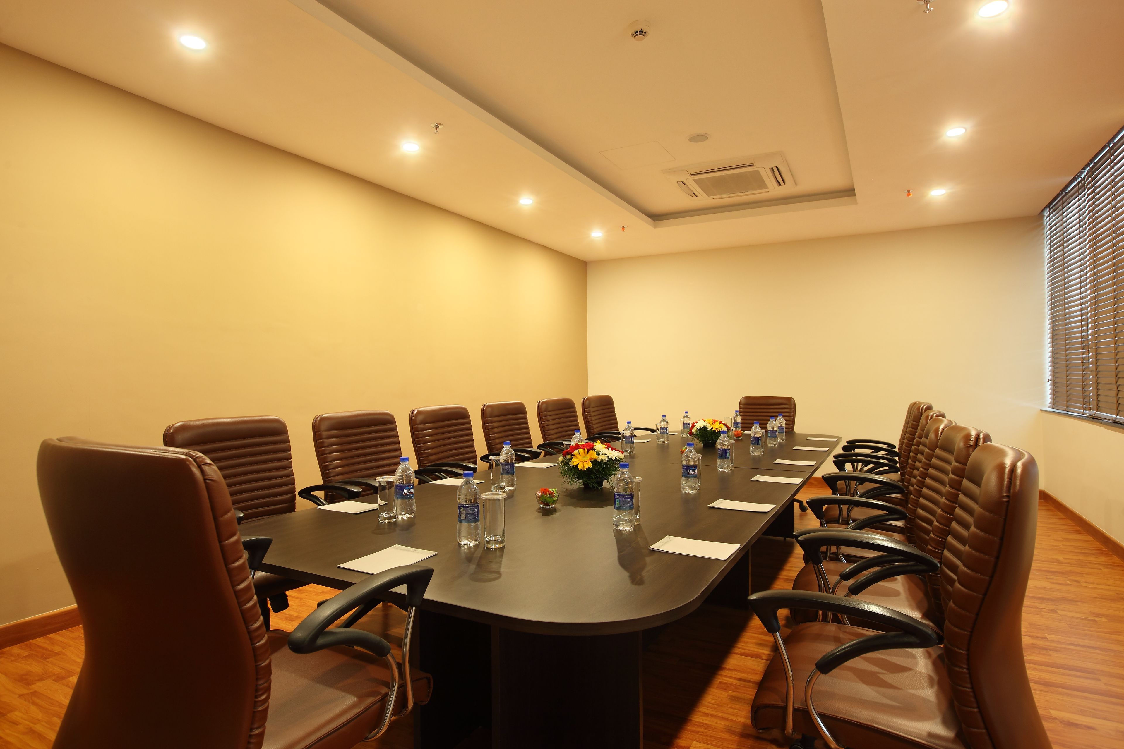 Un salón de conferencias con una larga mesa, sillas de cuero marrón, botellas de agua y un jarrón con flores.