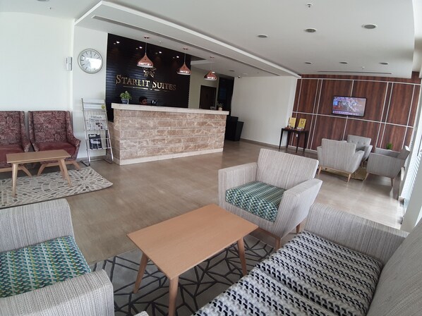 A hotel lobby with a reception desk, seating area, and a television screen displaying news.