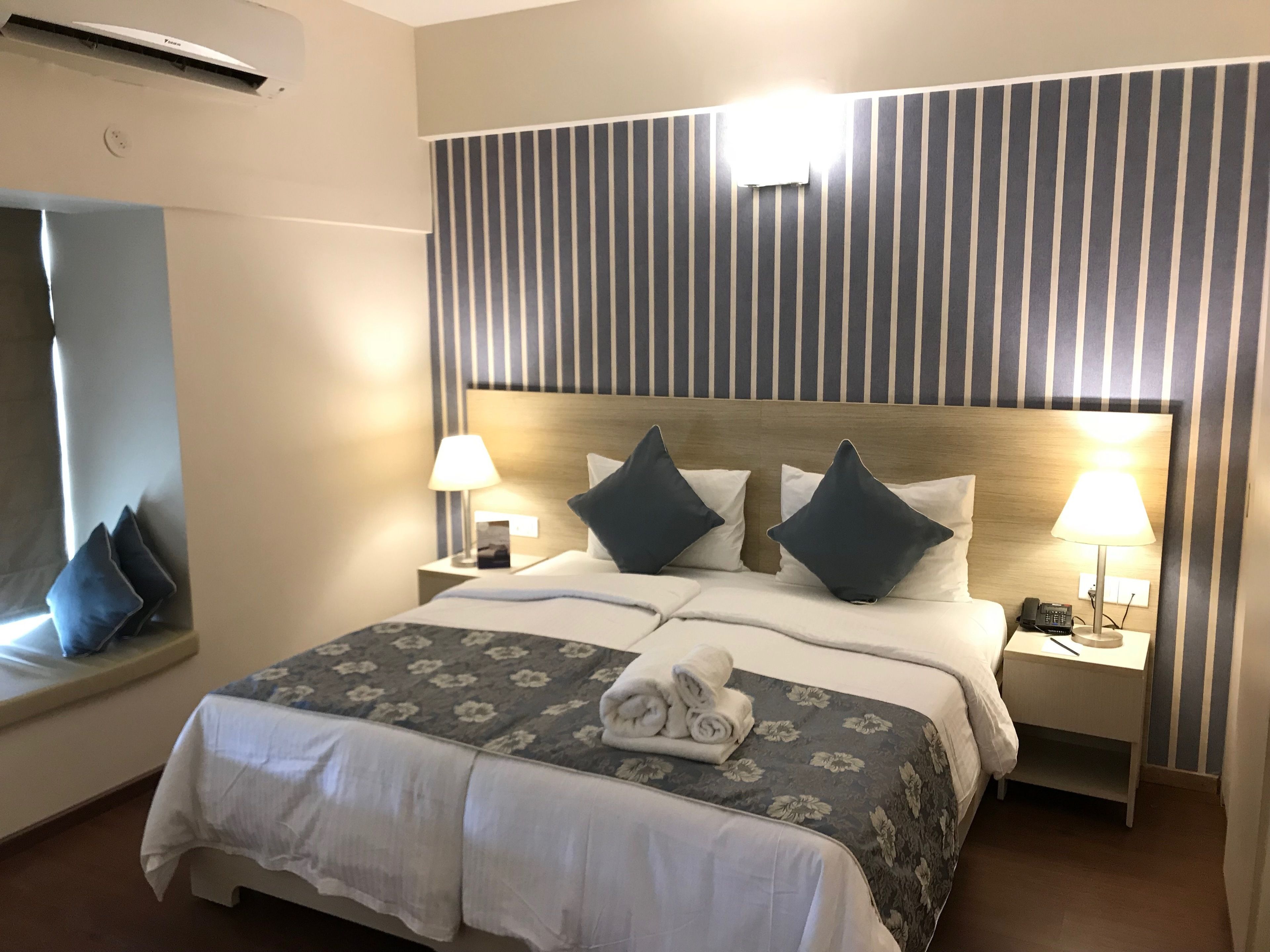 Habitació d'hotel amb un llit gran, dues taules de nit amb làmpades, un capçal de llit amb ratlles verticals i un aire condicionat fixat a la paret.