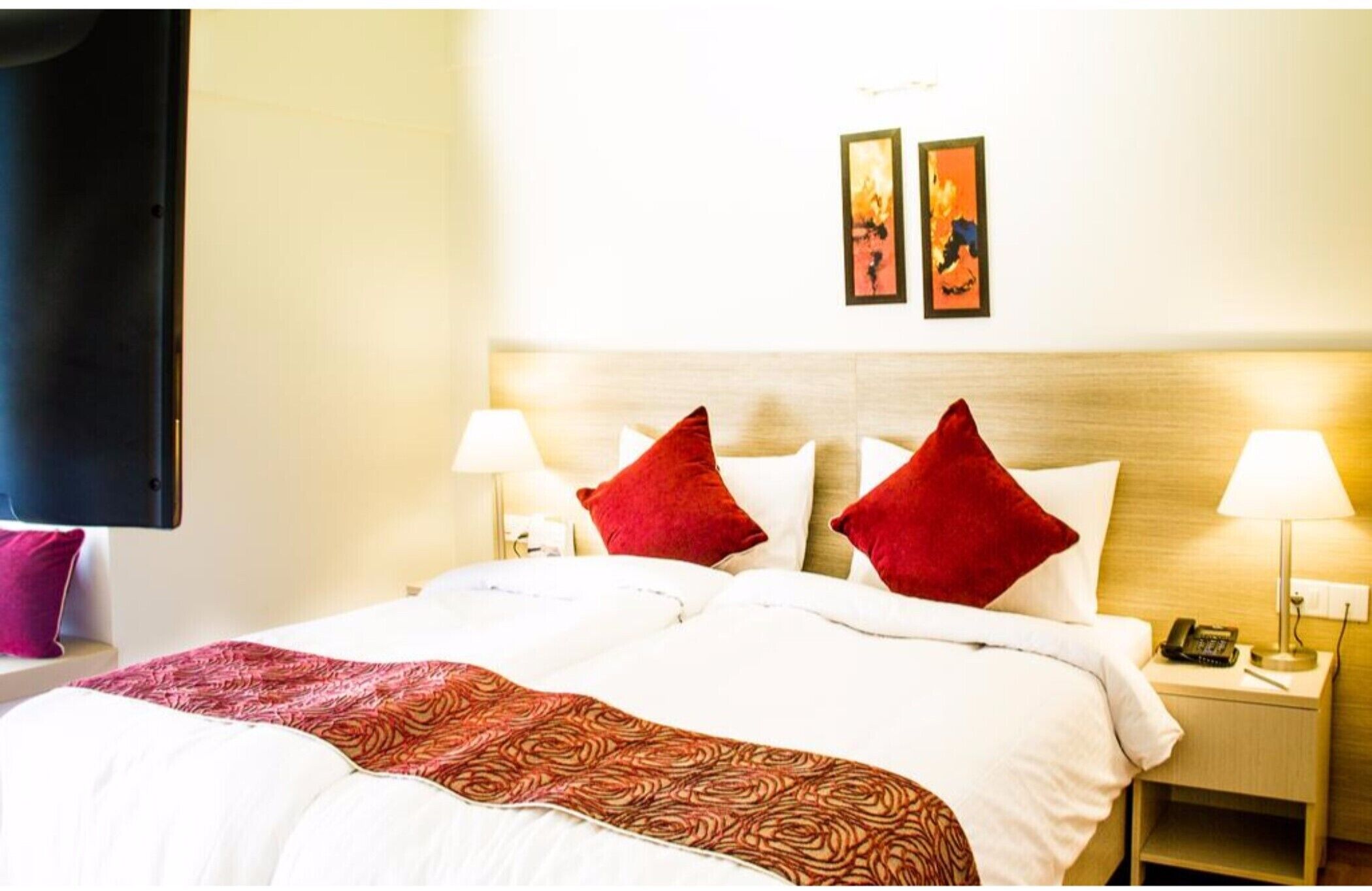Quarto de hotel com cama, duas luminárias de cabeceira, mesa de cabeceira com telefone e duas fotos emolduradas na parede.