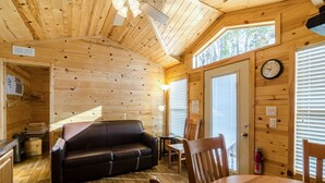 A cozy cabin interior with a wooden ceiling, a black leather sofa, a dining table with chairs, and a clock on the wall. - Wilderness Lakes RV Resort (Menifee)