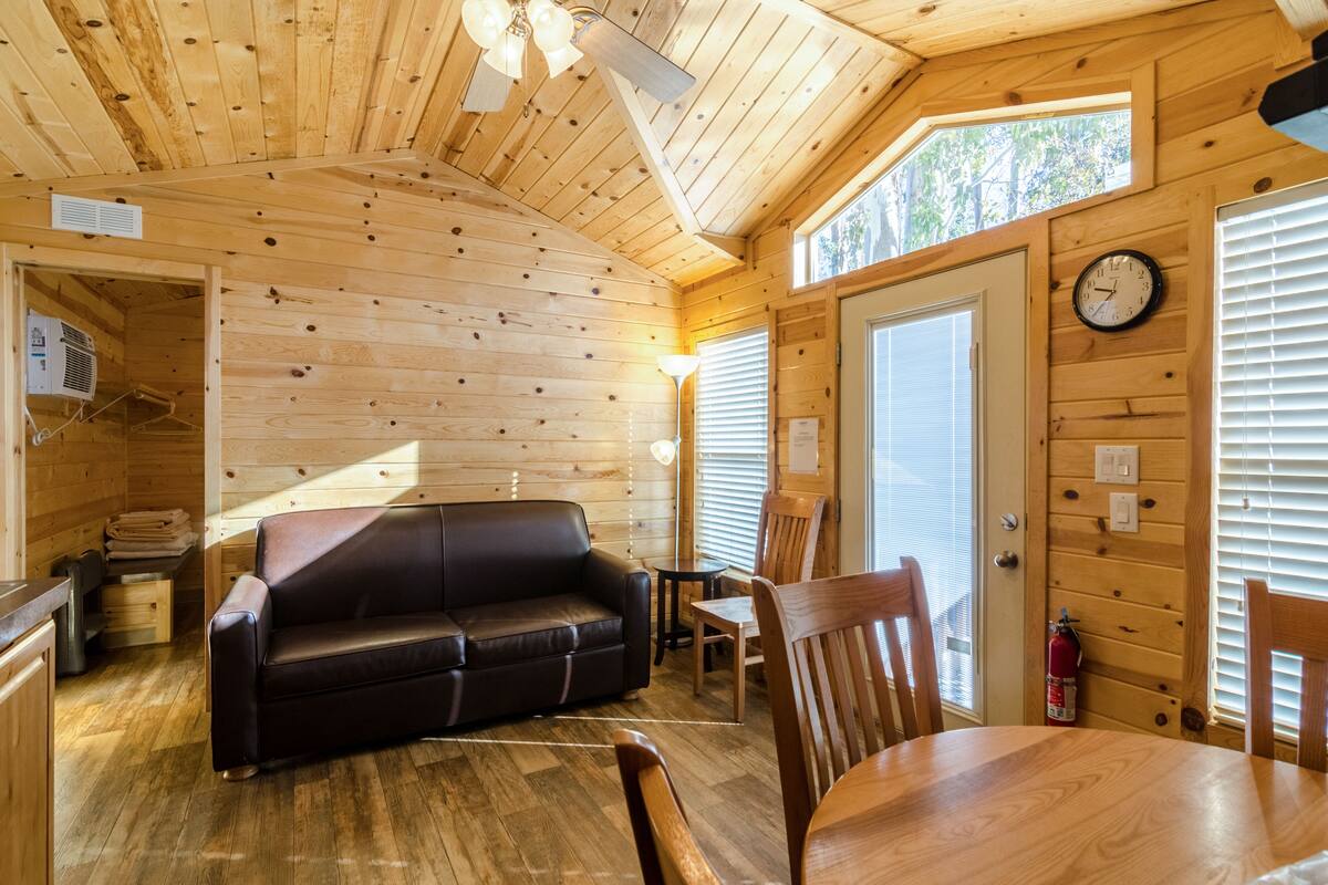 A cozy cabin interior with a wooden ceiling, a black leather sofa, a dining table with chairs, and a clock on the wall.