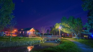 A nighttime scene with a small bridge over a pond, surrounded by trees and a lit building. - Wilderness Lakes RV Resort (Menifee)