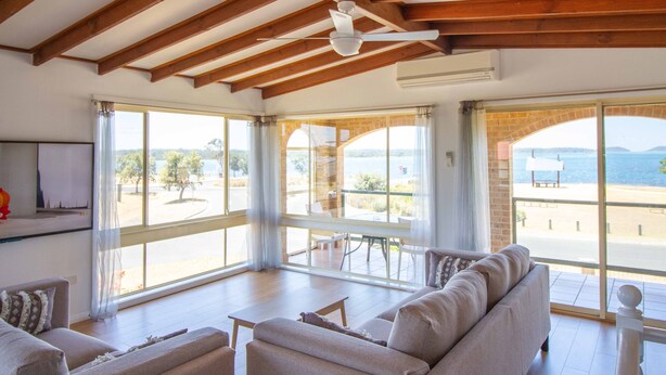 Un salón amplio con un sofá grande, una mesa de centro y un ventilador de techo. Grandes ventanas ofrecen vistas al exterior.