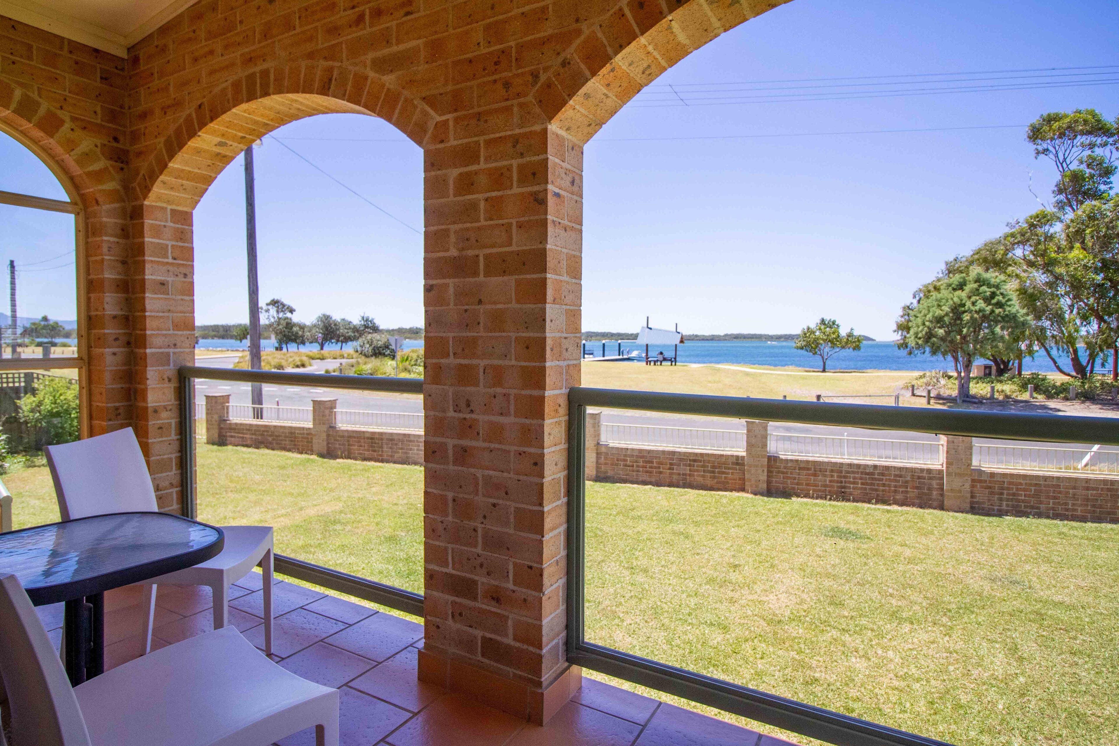 Ein Balkon mit Blick auf Strand und Meer, mit einer Ziegelstütze, weißen Stühlen und einem Glastisch.
