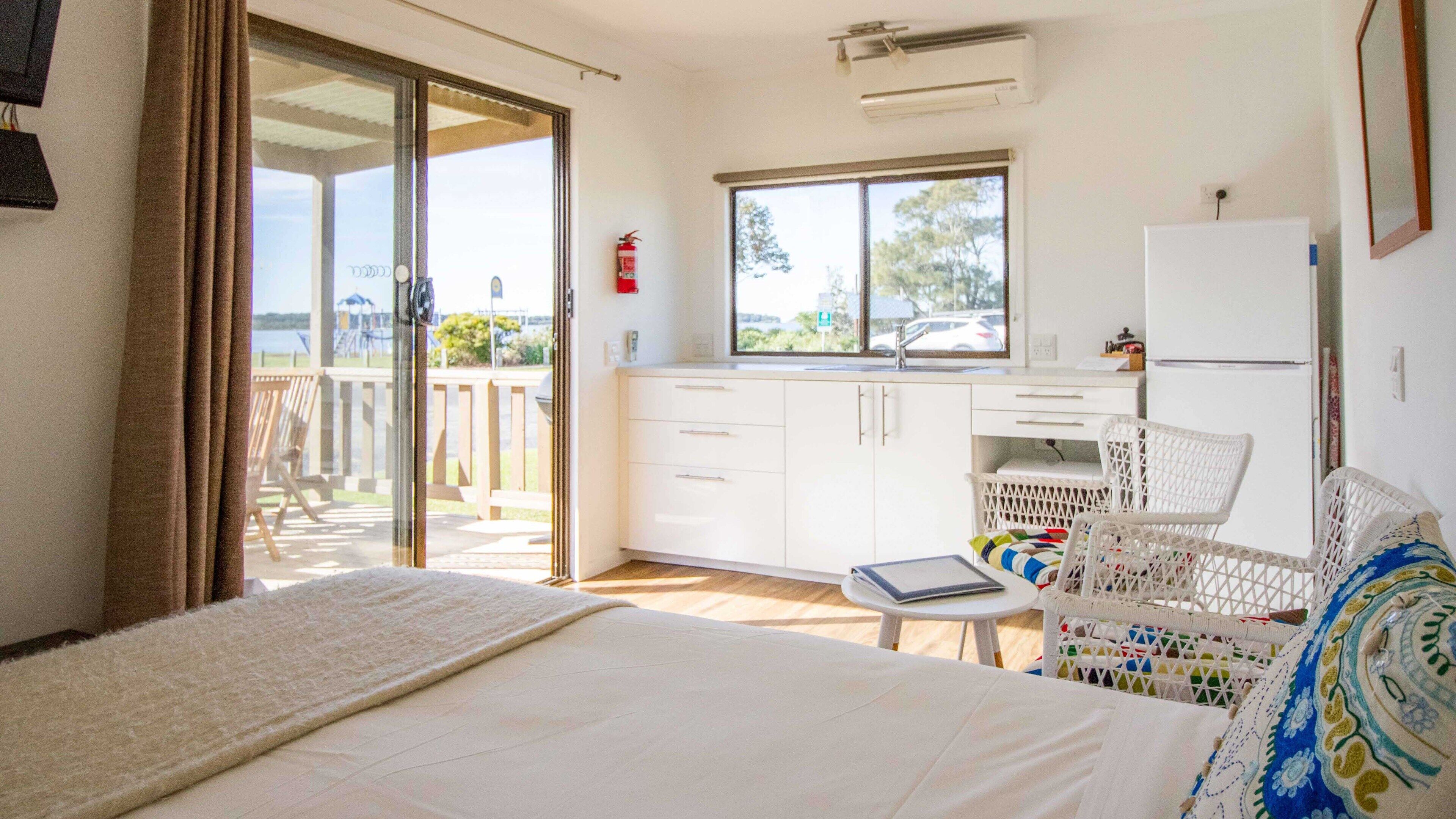 A room with a bed, a small table, a white wicker chair, a kitchenette, and a sliding glass door leading to an outdoor area.
