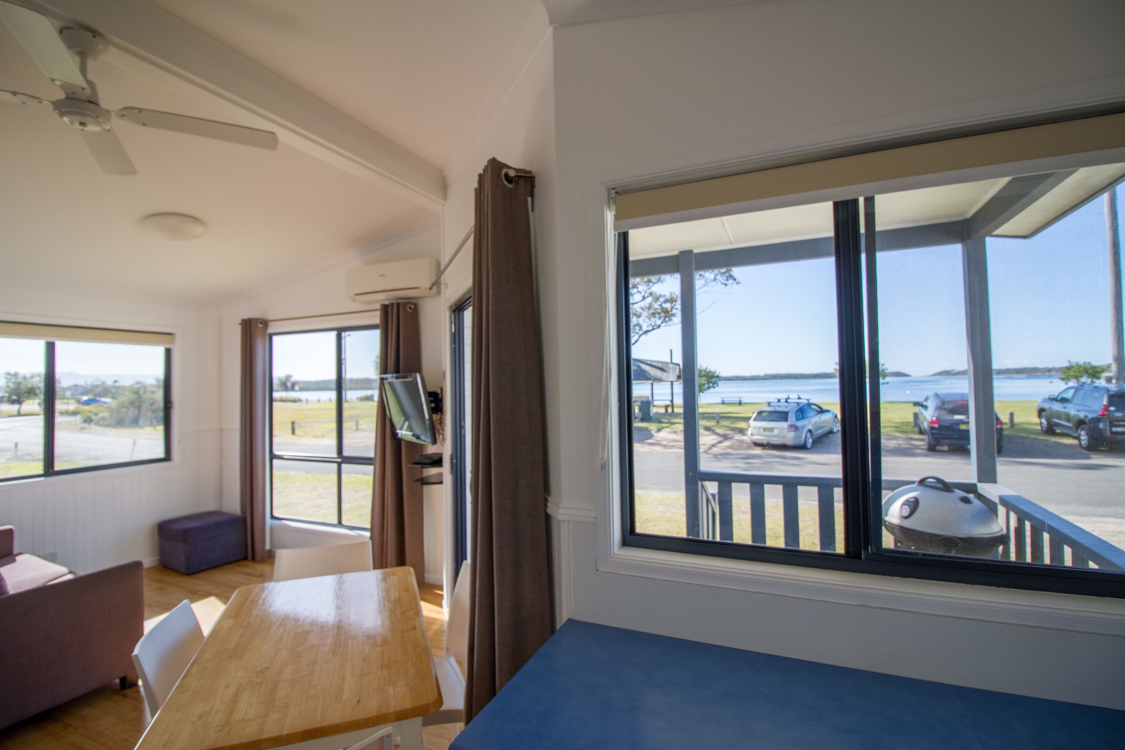 Una habitación con un gran ventanal, un ventilador de techo, una mesa de madera y un televisor.